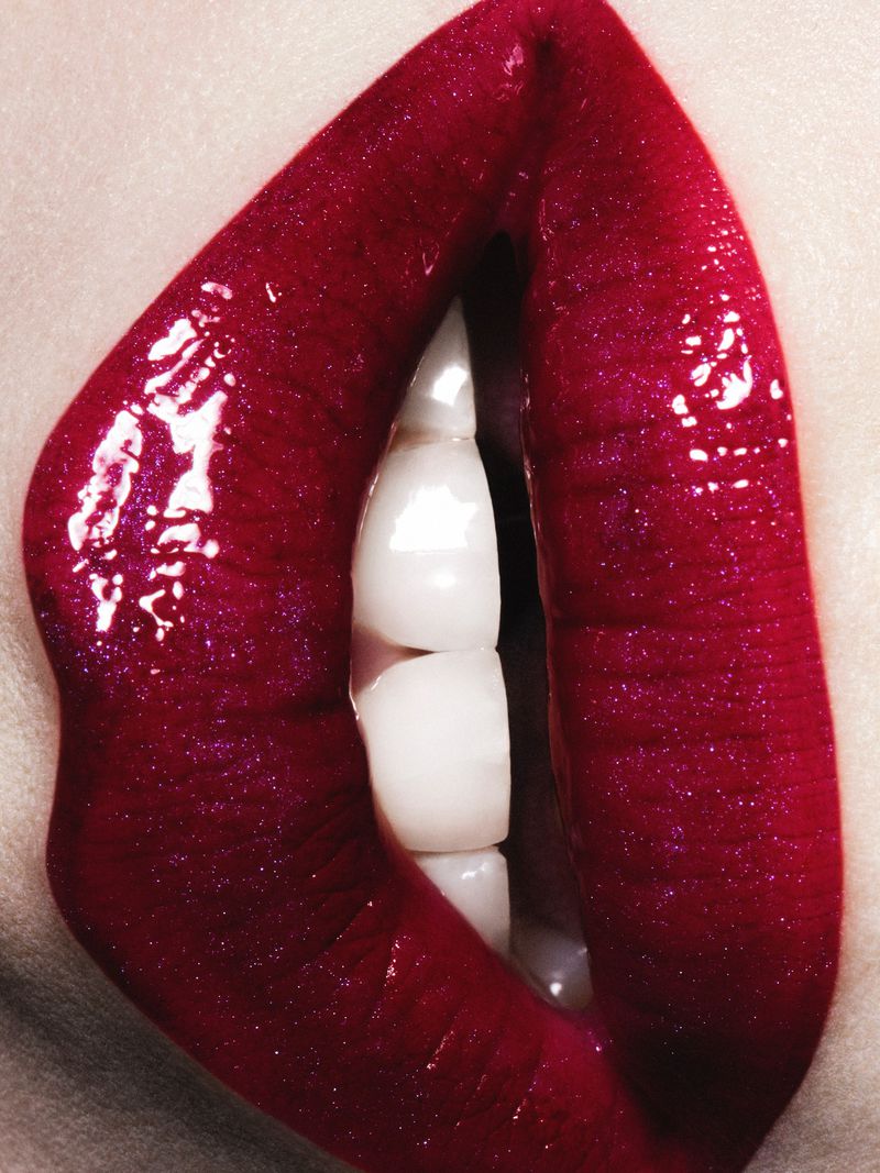 Close-up macro shot of glossy ruby red lipstick on lips with visible white teeth