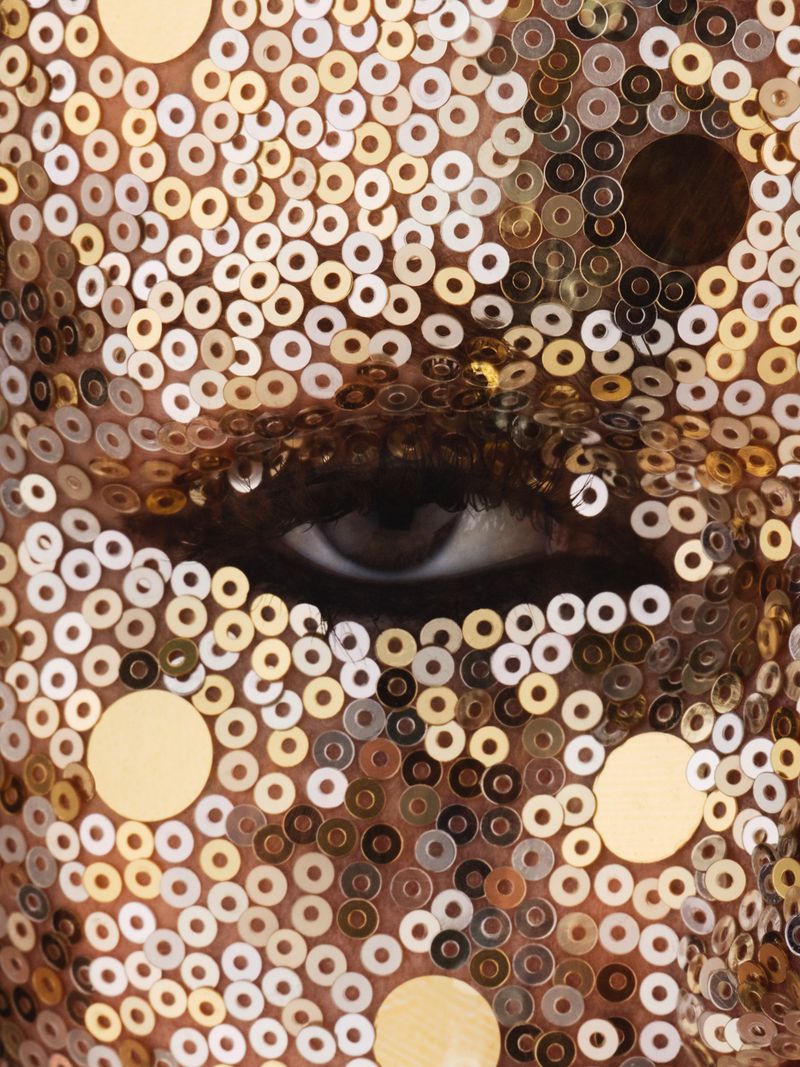 Close-up of a person's face covered in gold sequins, focusing on one eye with a serious expression in an indoor setting.