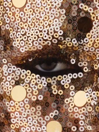 Close-up of a person's face covered in gold sequins, focusing on one eye with a serious expression in an indoor setting.