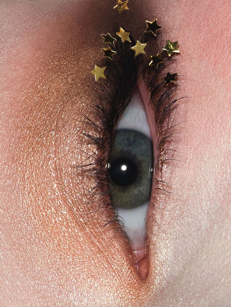 Close-up of a blue and green eye with gold star-shaped embellishments and gold shimmer eyeshadow for a beauty or fashion concept.