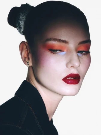 Woman with bold makeup: red lips, orange and blue eyeshadow, pink blush, wearing a black jacket and headscarf, set against white background.