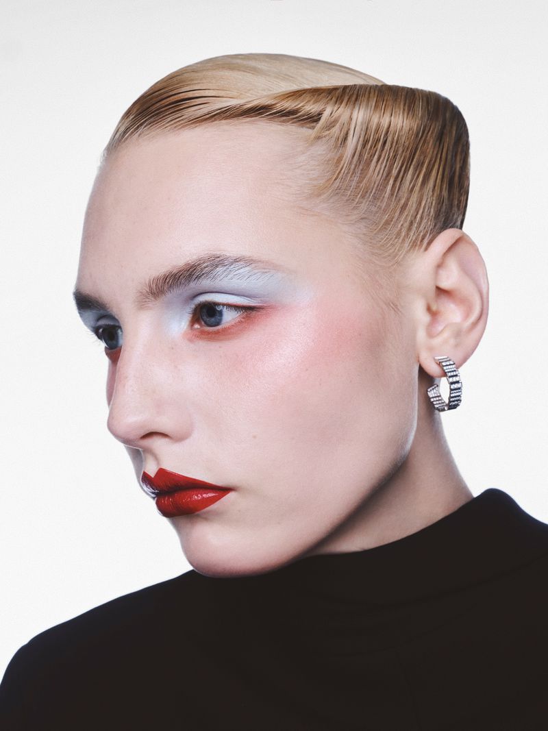 Close-up beauty shot of a blonde woman with red lipstick and blue eyeshadow, adorned with a silver earring, set against a bright white backdrop.