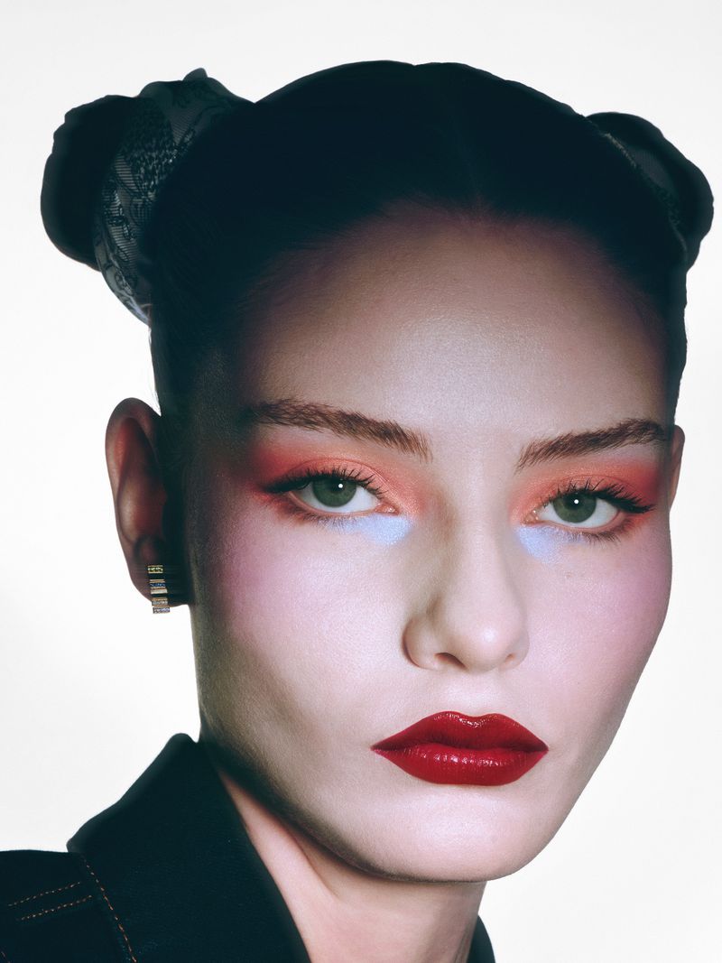 Close-up portrait of a young woman with dramatic makeup including red lipstick, orange eyeshadow, and face blush against a bright white background.