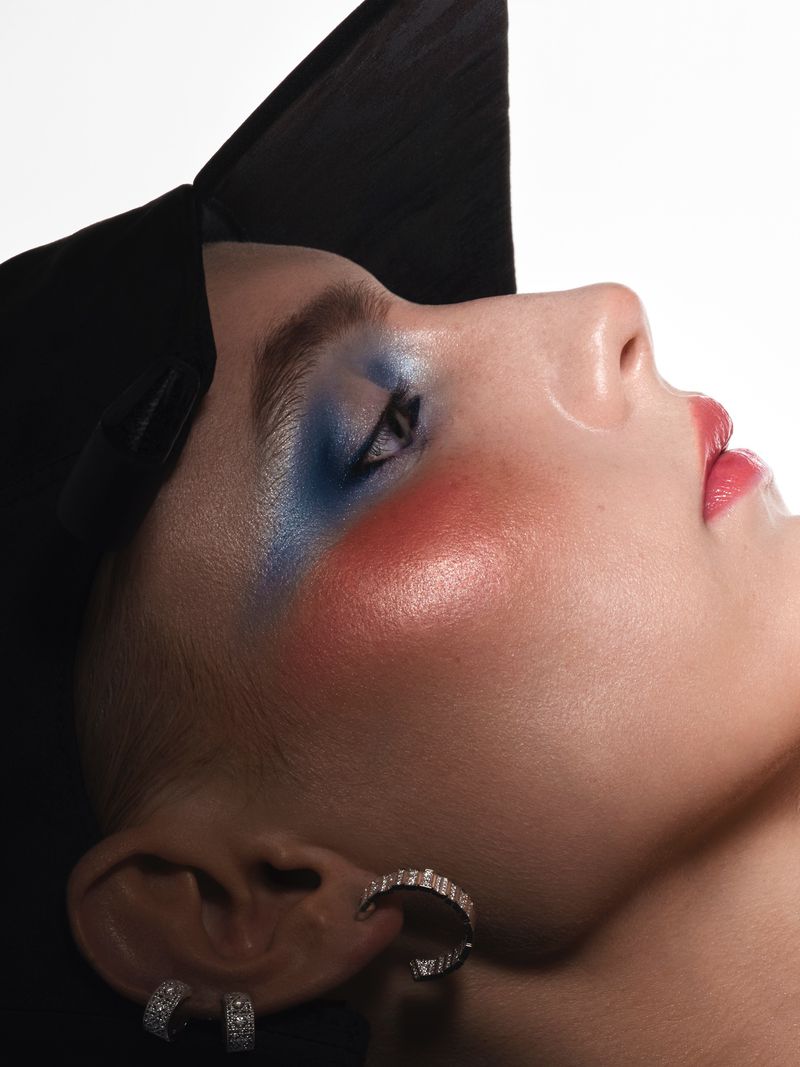 Close-up profile of a model wearing vibrant blue eyeshadow, pink blush, and a black hat against a white background.