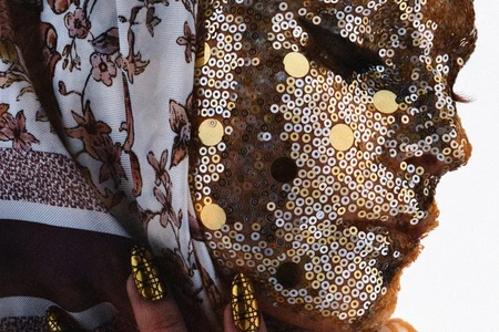 Close-up portrait of a model with gold sequins covering her face, wearing a floral patterned headscarf and metallic gold manicured nails.