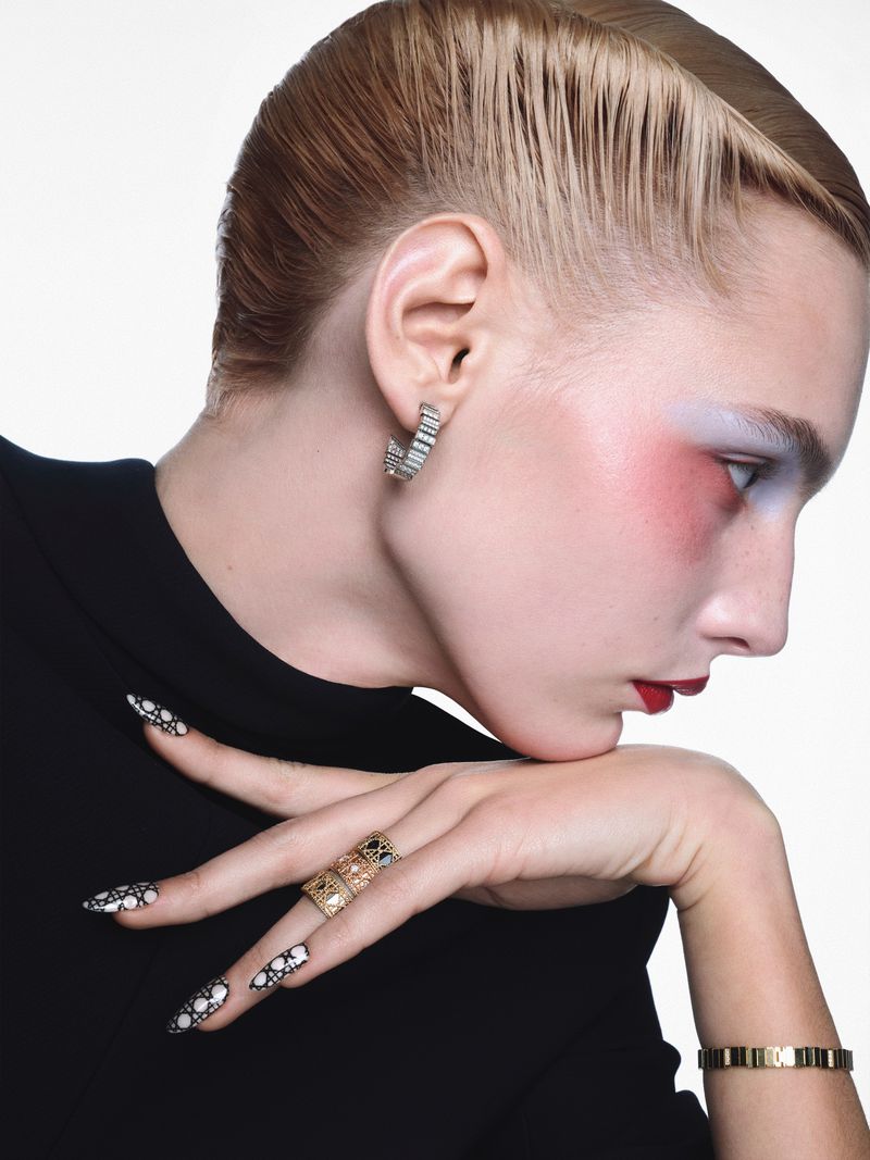Close-up profile of a model with artistic eye makeup, intricate patterned nails, and gold rings against a white background