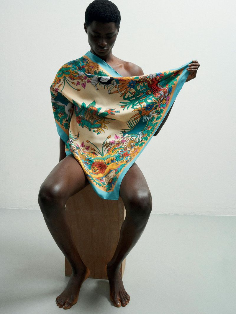 A dark-skinned person sits on a wooden block draped with a colorful patterned silk scarf in a studio setting with a white backdrop.