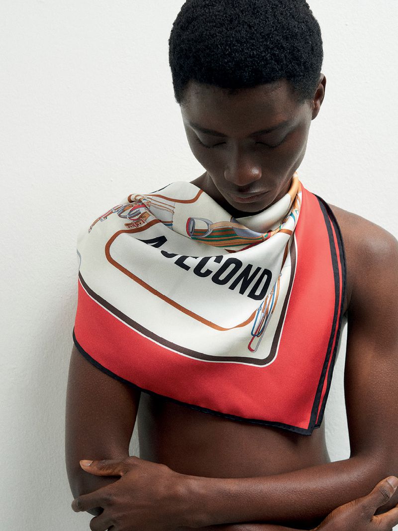 Close-up of a shirtless person with dark hair and skin, a printed scarf draped across their shoulders, and standing against a white wall.