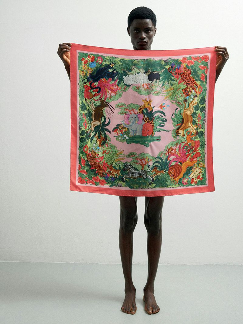 An adult male stands in a photo studio holding a large, patterned pink scarf with jungle animal imagery that covers his torso.