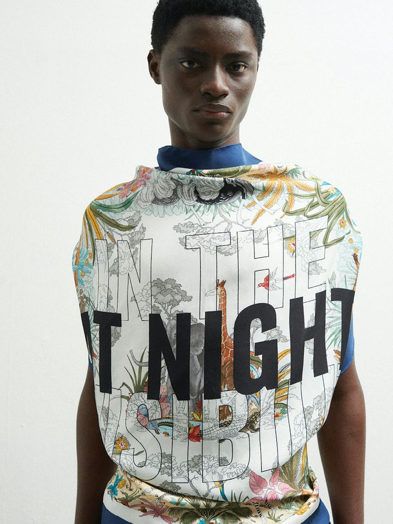 Person with dark complexion wearing a designer scarf with illustration and large text on a white backdrop in an indoor photo studio.