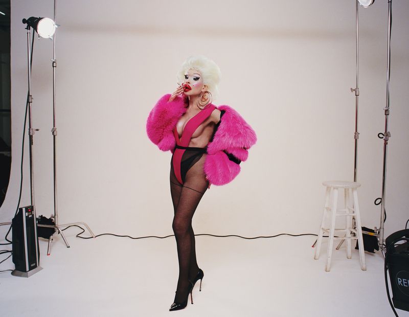Drag queen in pink faux fur coat posing in a photo studio with white backdrop, lighting equipment, and a stool visible.