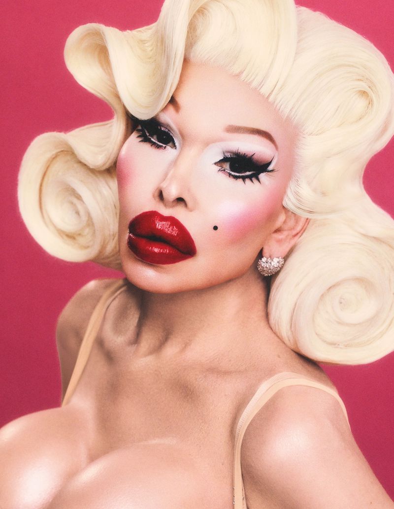 Glamorous portrait of a person with platinum blonde hair, red lips, and heavy makeup against a bright pink backdrop in a photo studio.