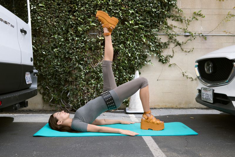 Woman in grey workout clothes doing yoga bridge pose with one leg up wearing platform shoes on a teal mat in a parking lot.