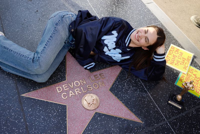 Smiling brunette woman lies on Hollywood Walk of Fame star dedicated to Devon Lee Carlson, wearing jeans and a varsity jacket.