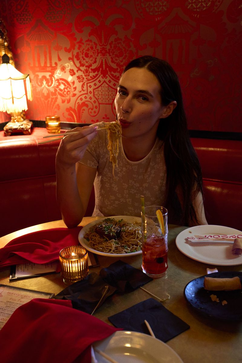 Young woman with long dark hair is at a restaurant table, eating noodles with chopsticks. Red damask wallpaper in background.