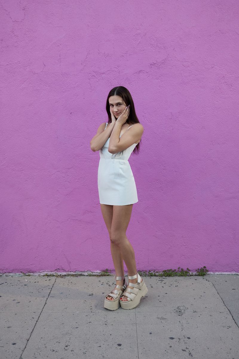 A full shot of a woman in a white dress and platform sandals posing in front of a purple wall on a sidewalk in natural light.