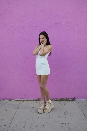 A full shot of a woman in a white dress and platform sandals posing in front of a purple wall on a sidewalk in natural light.