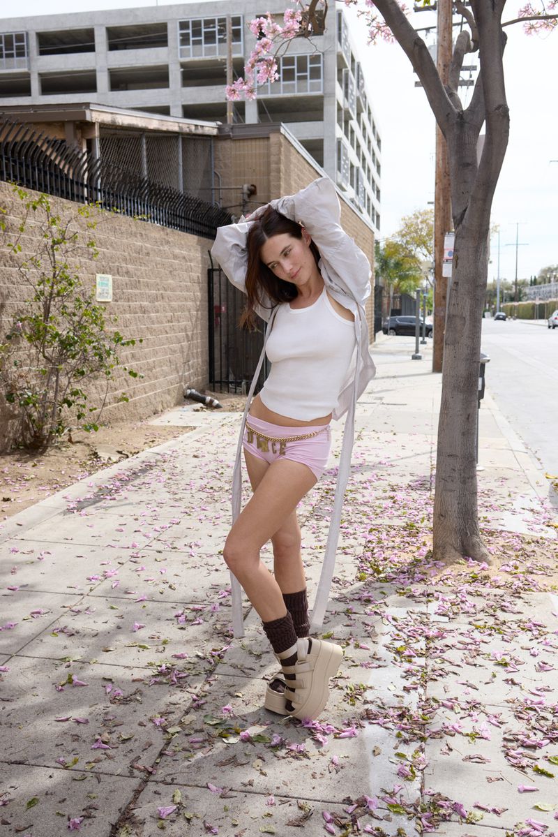 Woman in pink shorts, white tank top, platform shoes and jacket standing in front of city building with a tree with pink flowers.