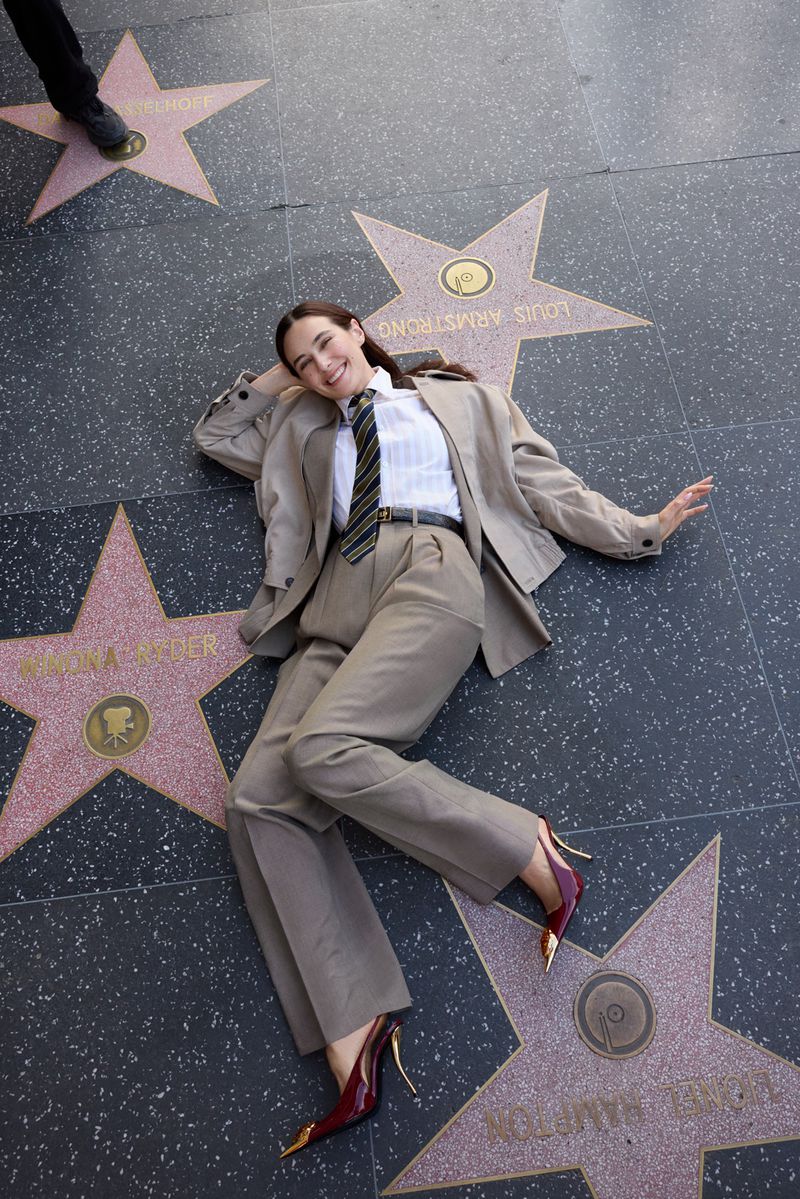 Woman in a beige suit lies on the Hollywood Walk of Fame with star-shaped plaques. Her burgundy high heels have gold accents.