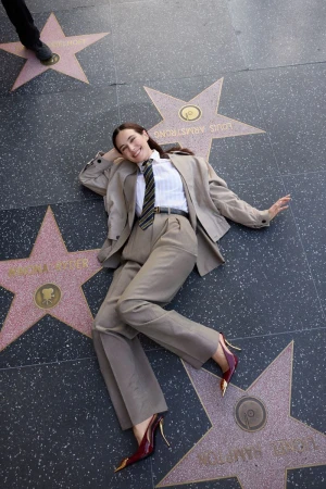 Woman in a beige suit lies on the Hollywood Walk of Fame with star-shaped plaques. Her burgundy high heels have gold accents.