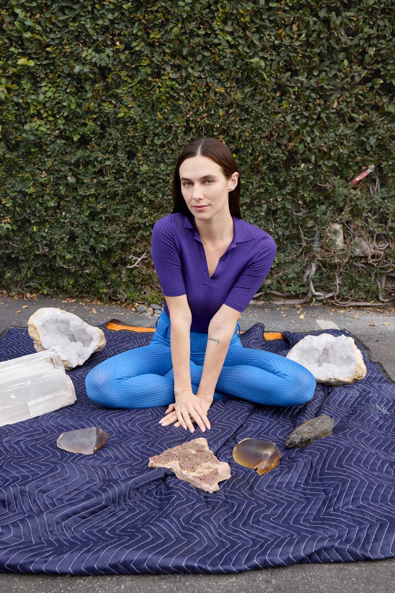 A woman poses cross-legged on a blue quilt, surrounded by crystals, against a green leafy backdrop in natural, eye-level outdoor shot.