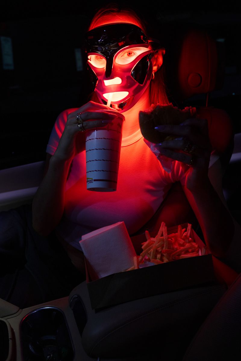 Person in a futuristic mask eats a burger and fries in car with red lighting, holding a drink with straw. Fast food meal.