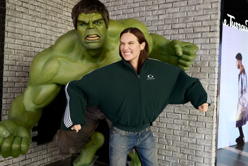 Woman posing next to the Incredible Hulk statue, wearing a cropped green jacket and jeans, smiling with a brick wall background.