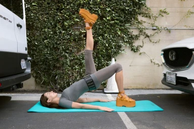 Devon Lee Carlson lying on a turquoise yoga mat doing a bridge pose outdoors wearing platform sandals