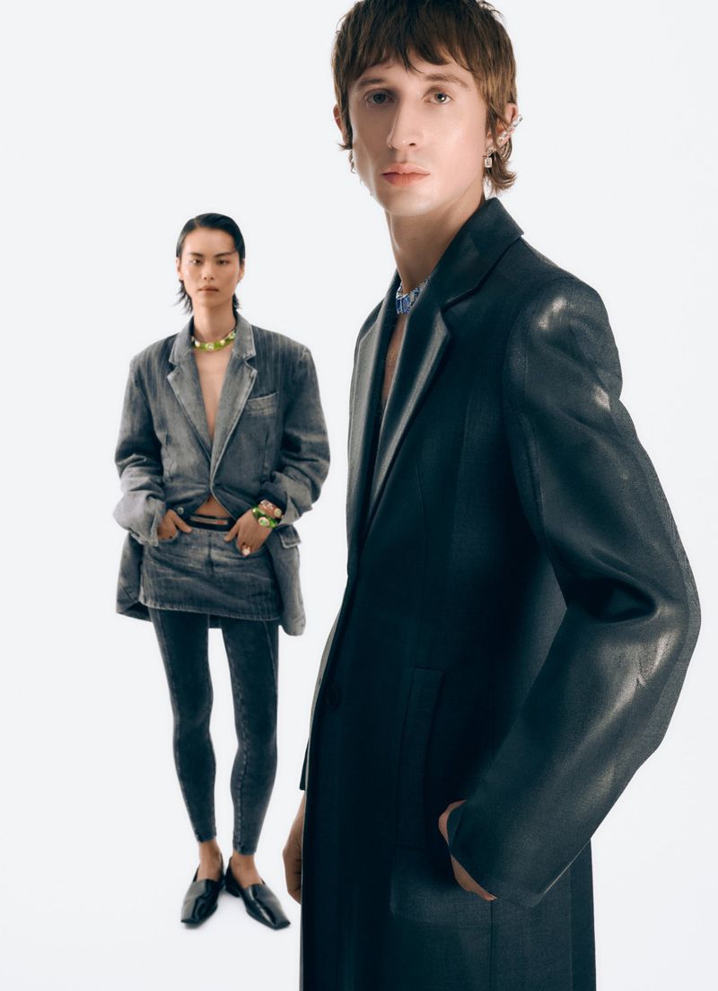 Two fashion models in a studio setting. A woman wears a denim suit and the man wears a black leather suit, posing against a white background.