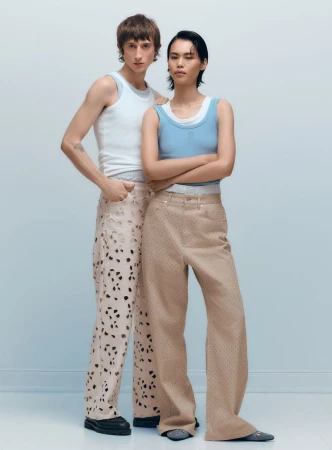 Two fashion models in a studio setting, the male with a white tank and perforated pants, and the female with a blue tank and tan pants.