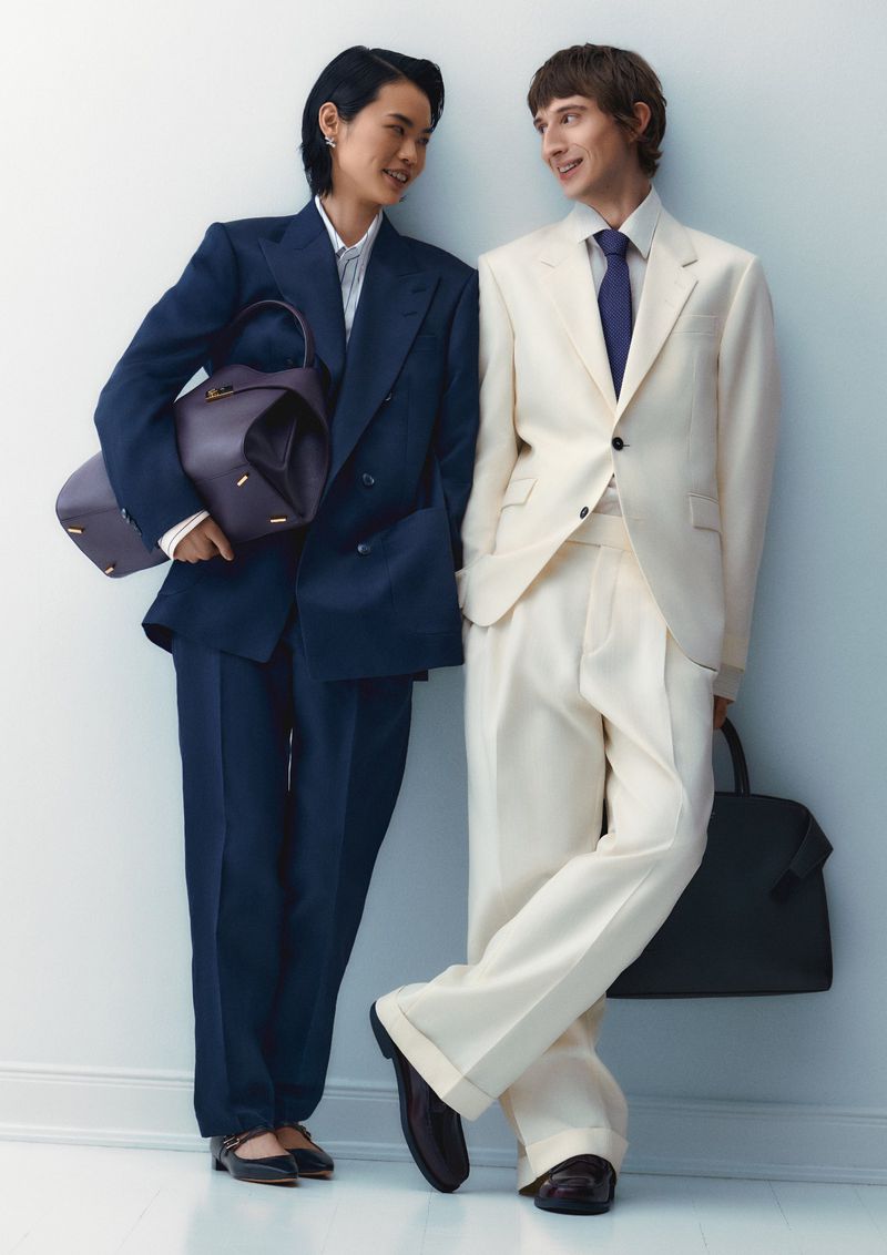 Two people, a man and a woman, in cream and navy suits stand close together. The woman carries a purple bag, and the man a black bag.