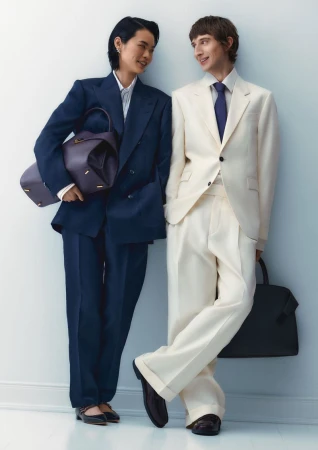 Two people, a man and a woman, in cream and navy suits stand close together. The woman carries a purple bag, and the man a black bag.