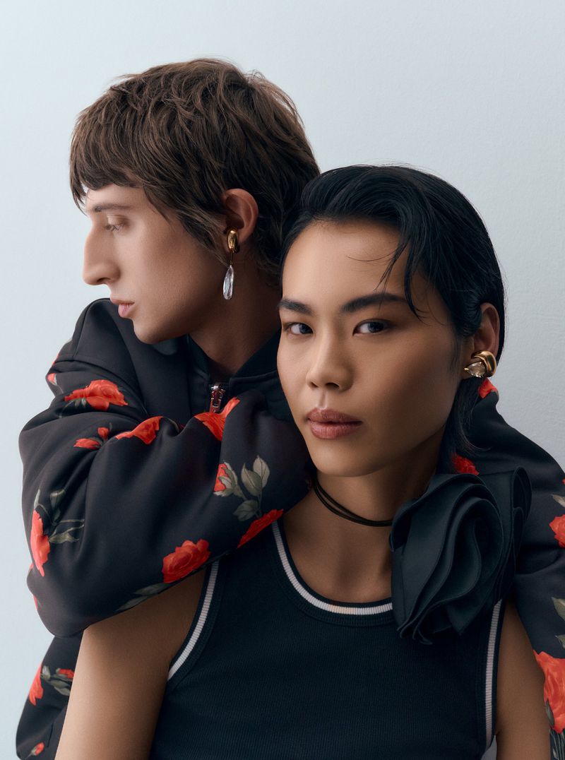 Two models pose indoors; one with arms around the other, showcasing earrings. Both are wearing jackets and tank tops with neutral background.