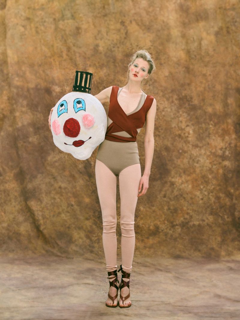 Woman in studio holding plush clown head with painted face, wearing brown top, tan bottoms, and gladiator sandals on a brown backdrop.