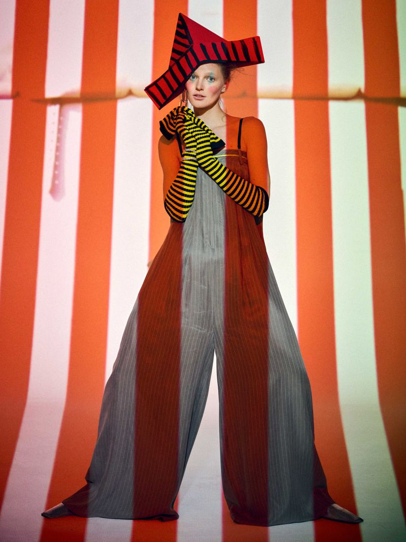 Model poses in a striped jumpsuit and a hat with red and black stripes, standing against an orange and white striped backdrop.