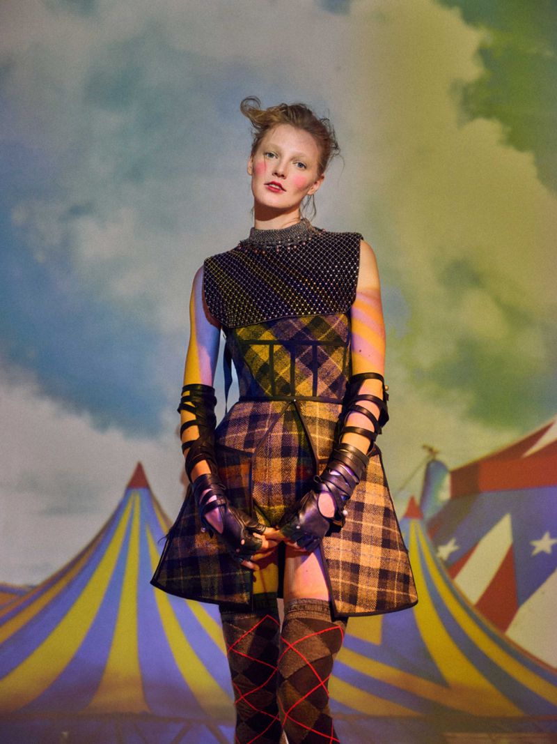Blond female model in patterned mini dress, black opera gloves, and knee socks, posing in front of a circus tent against a cloudy sky.