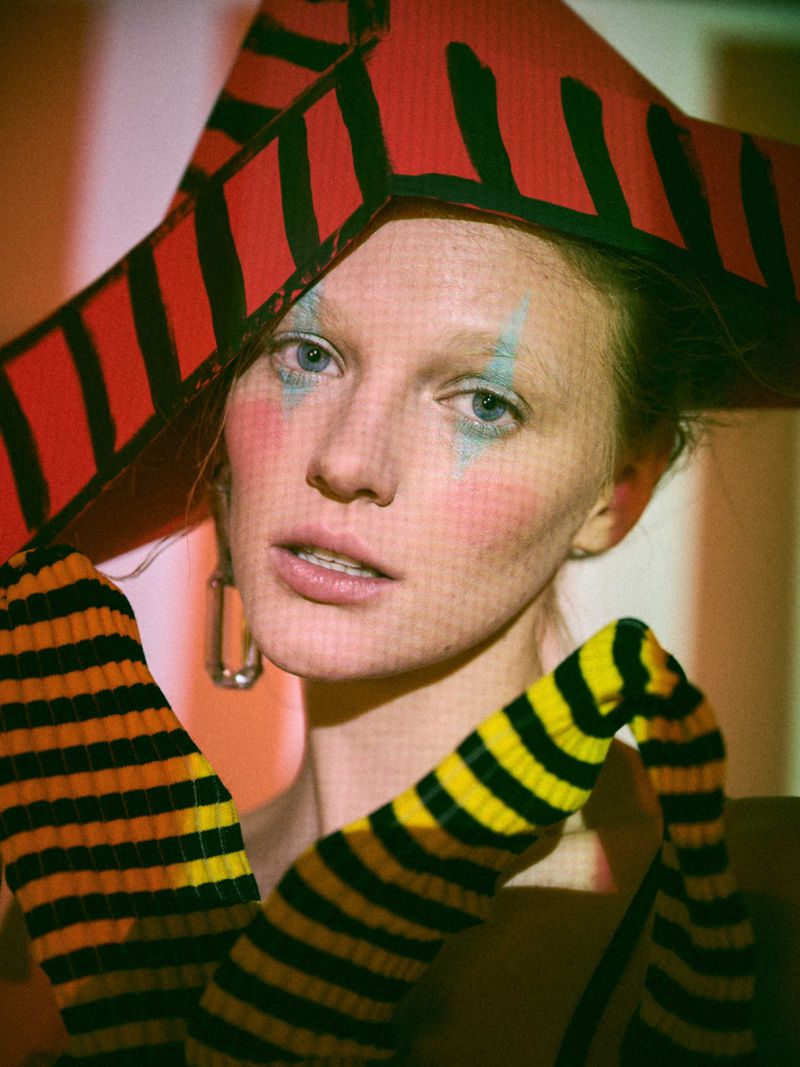 Portrait of a model in a striped hat with artistic makeup. The image features a close-up shot with orange and yellow tones.