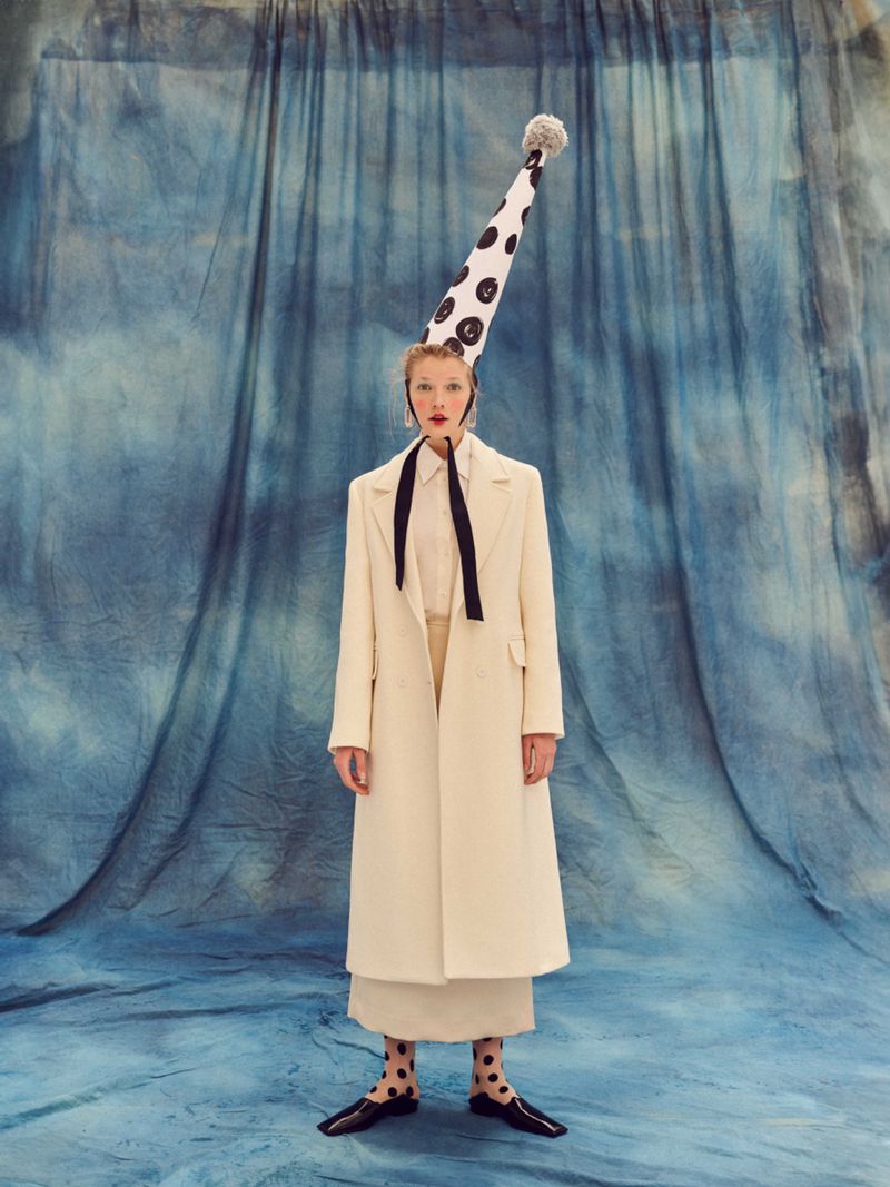 A woman in a cream-colored coat poses against a blue textured backdrop wearing a polka dot hat, matching socks and patent shoes.