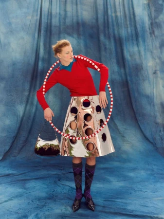 Fashion model poses with a red and white hula hoop wearing a shiny silver skirt with black and gray patterned socks in a studio.