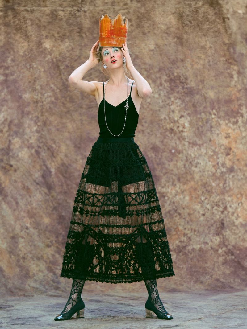 Woman in black dress and paper crown gazes upward against a mottled background. Full shot in a studio with dramatic lighting.