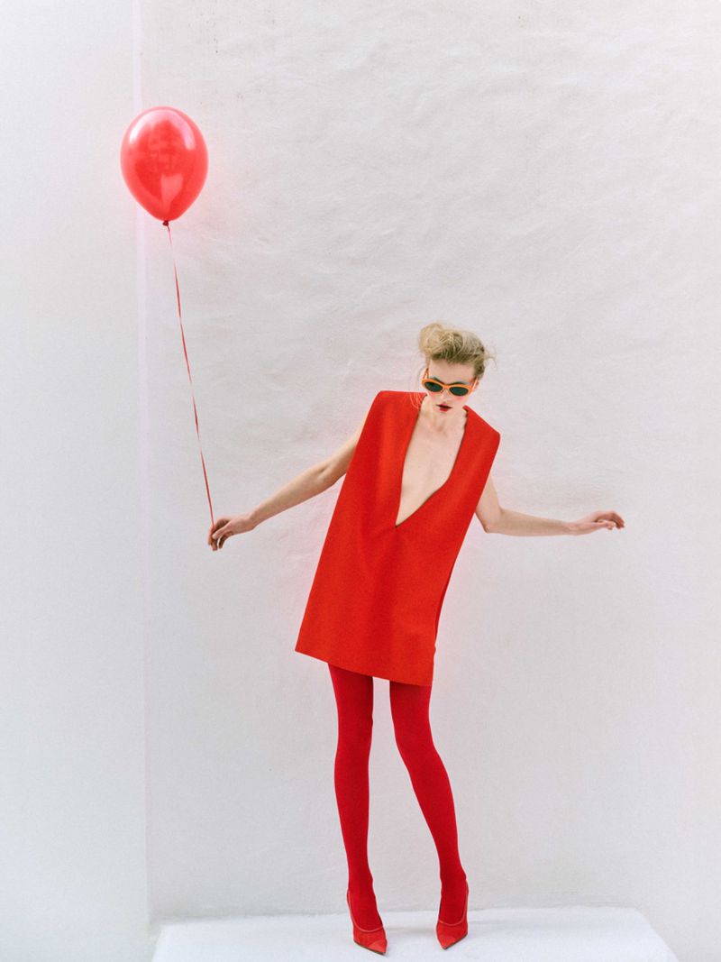 A tall blonde woman in a red dress and matching tights and heels poses with a red balloon in a white studio setting.