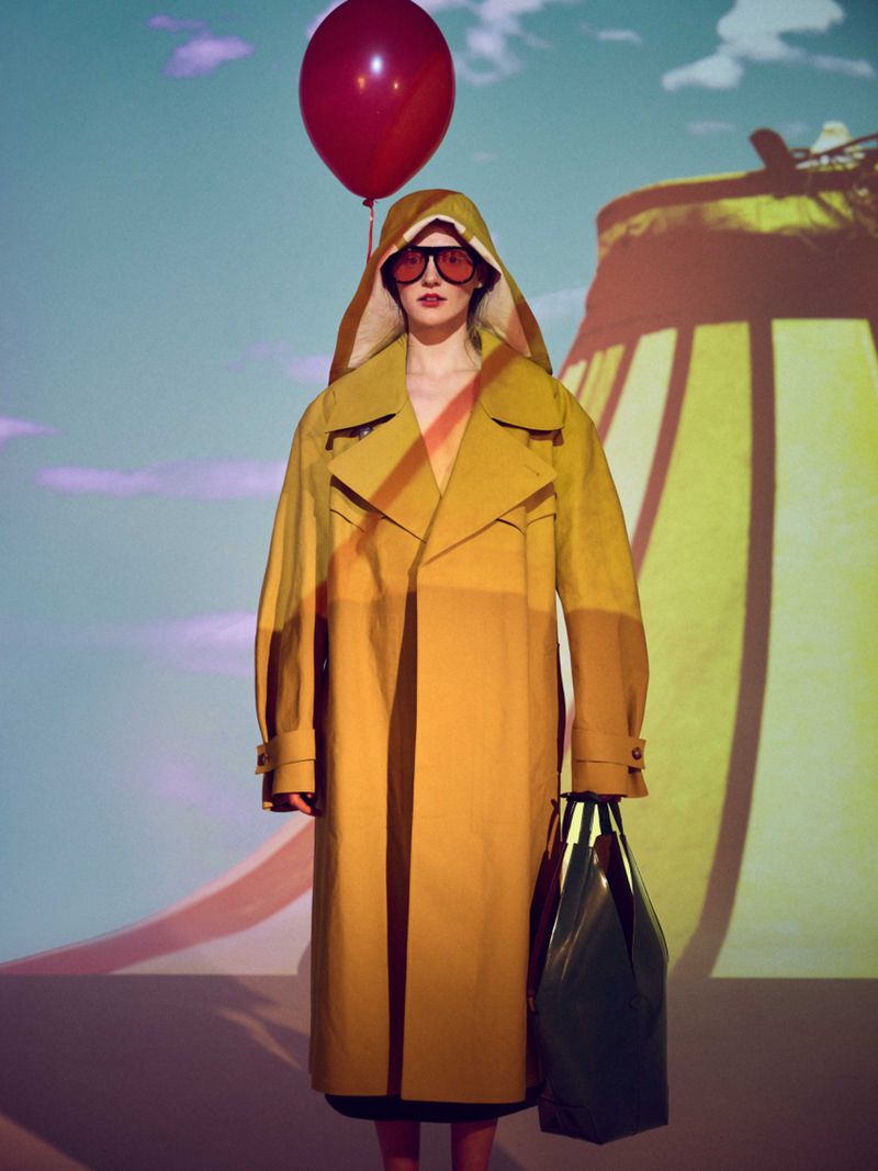 A stylish woman in a yellow trench coat and red sunglasses poses with a balloon against a surreal backdrop in a fashion studio.