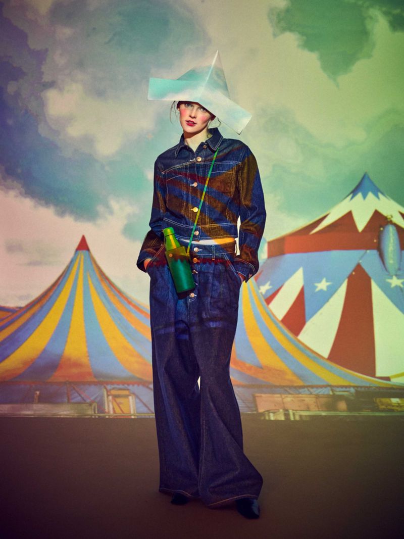 Full-length shot of a woman in a denim outfit and hat, holding a green bottle, against a backdrop of a circus tent and clouds.