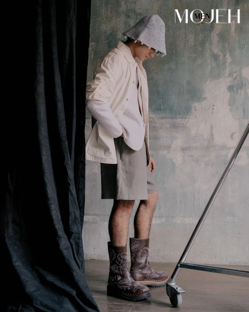 Studio shot of an adult male in a neutral-toned outfit with a knit hat and brown boots, posing against a textured backdrop with draped fabric.