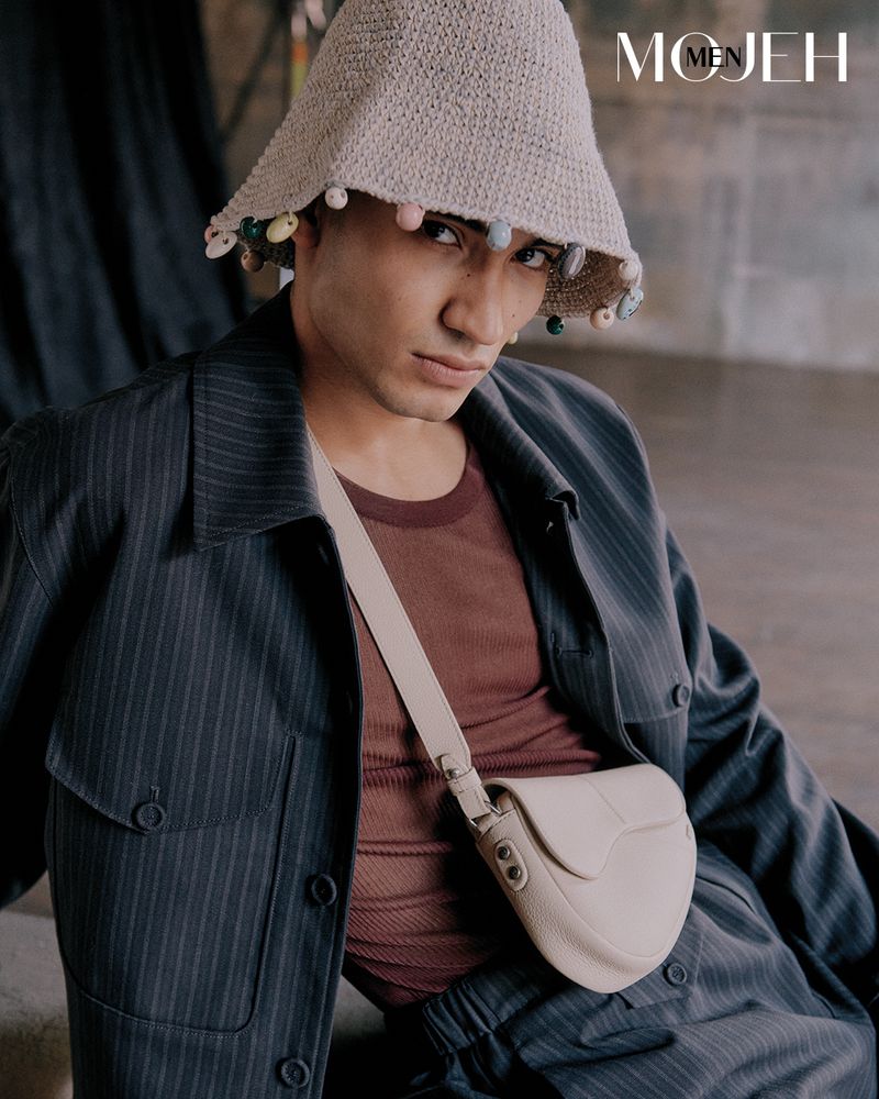 Stylish man in pinstripe suit and bucket hat poses with a cream crossbody bag, showcasing a fashion-forward, neutral aesthetic.