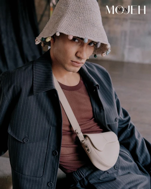Stylish man in pinstripe suit and bucket hat poses with a cream crossbody bag, showcasing a fashion-forward, neutral aesthetic.