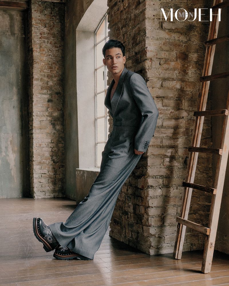 Model in grey suit leaning against exposed brick wall next to a wooden ladder