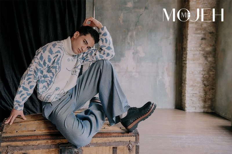 A stylish adult male poses while seated on a vintage trunk indoors wearing a patterned sweater, pants, and shoes against a textured wall.