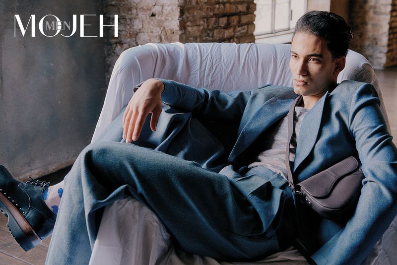 Adult male reclining in a chair, wearing a blue suit and carrying a handbag, in a brick-walled interior space. 