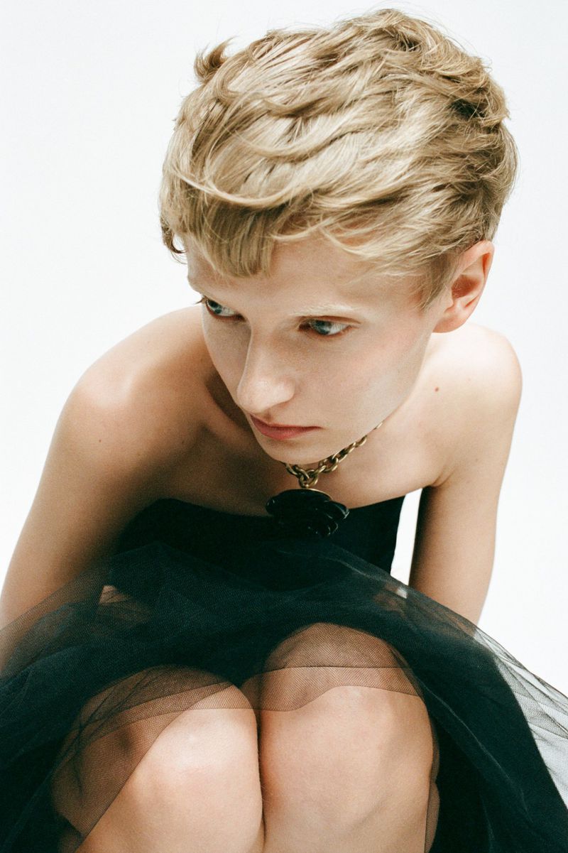 A young fair-skinned person poses in a strapless black dress against a bright white background in a studio portrait shot.